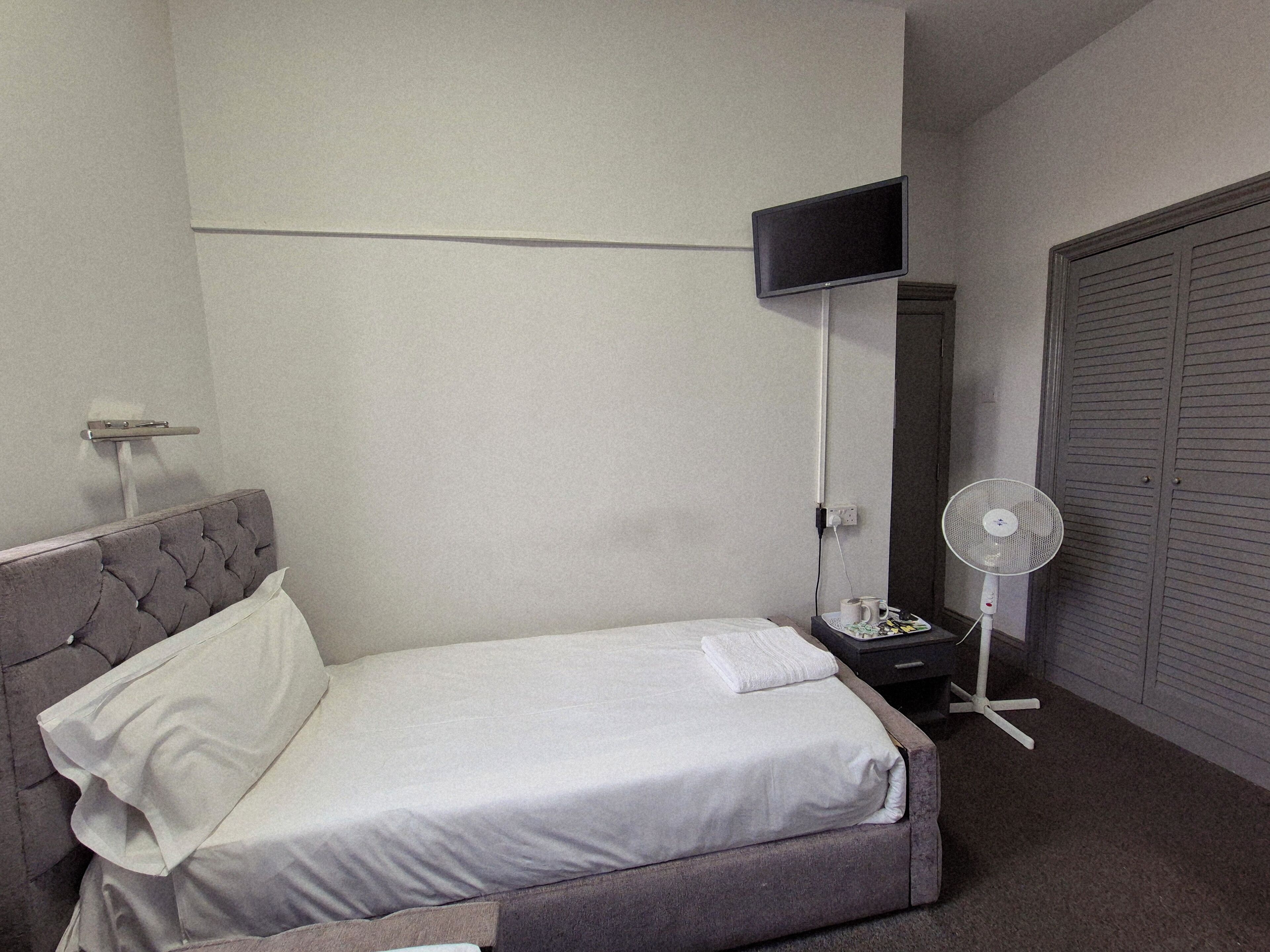 A neatly made bed with a grey headboard, a bedside table with a lamp, a fan, and a closed wardrobe.