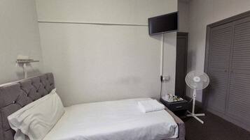 Un lit bien fait, avec une tête de lit grise, une table de chevet avec une lampe, un ventilateur et une armoire fermée.