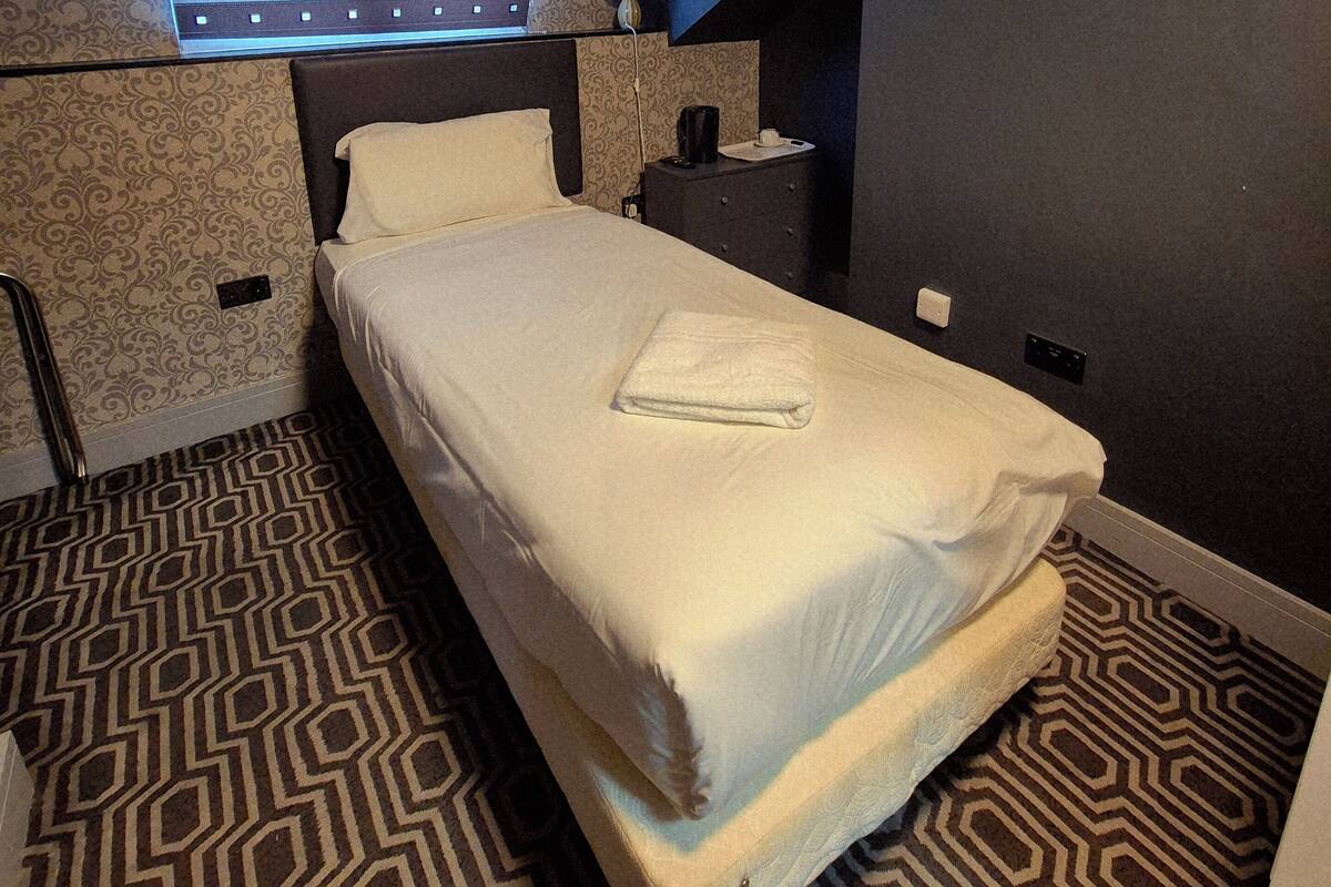 A hotel room with a bed, a nightstand, a lamp, a window, and a patterned carpet.