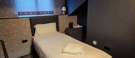Una habitación de hotel con una cama, una mesita de noche, una lámpara, una ventana y una alfombra estampada.