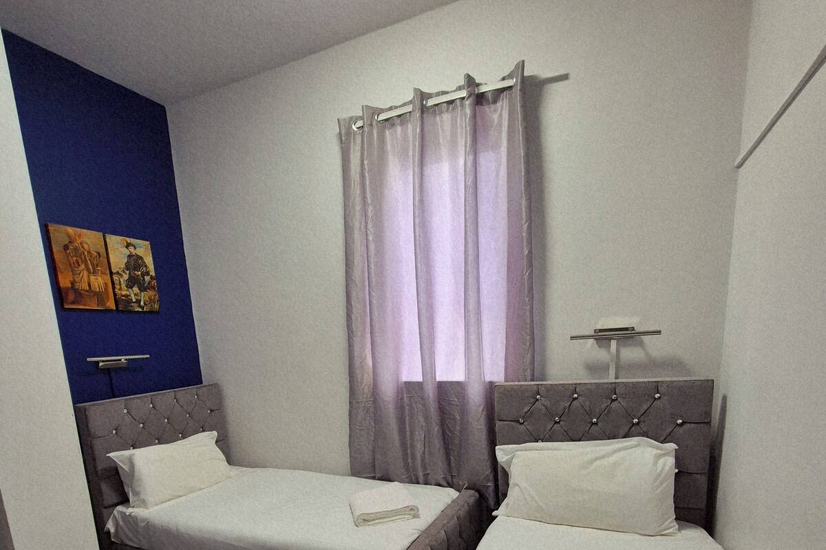 A hotel room with two single beds, a small bedside table, a nightstand, a wall-mounted clock, and a window with sheer curtains.