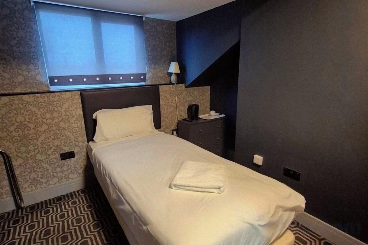 A hotel room with a bed, a nightstand, a lamp, and a window with blinds.
