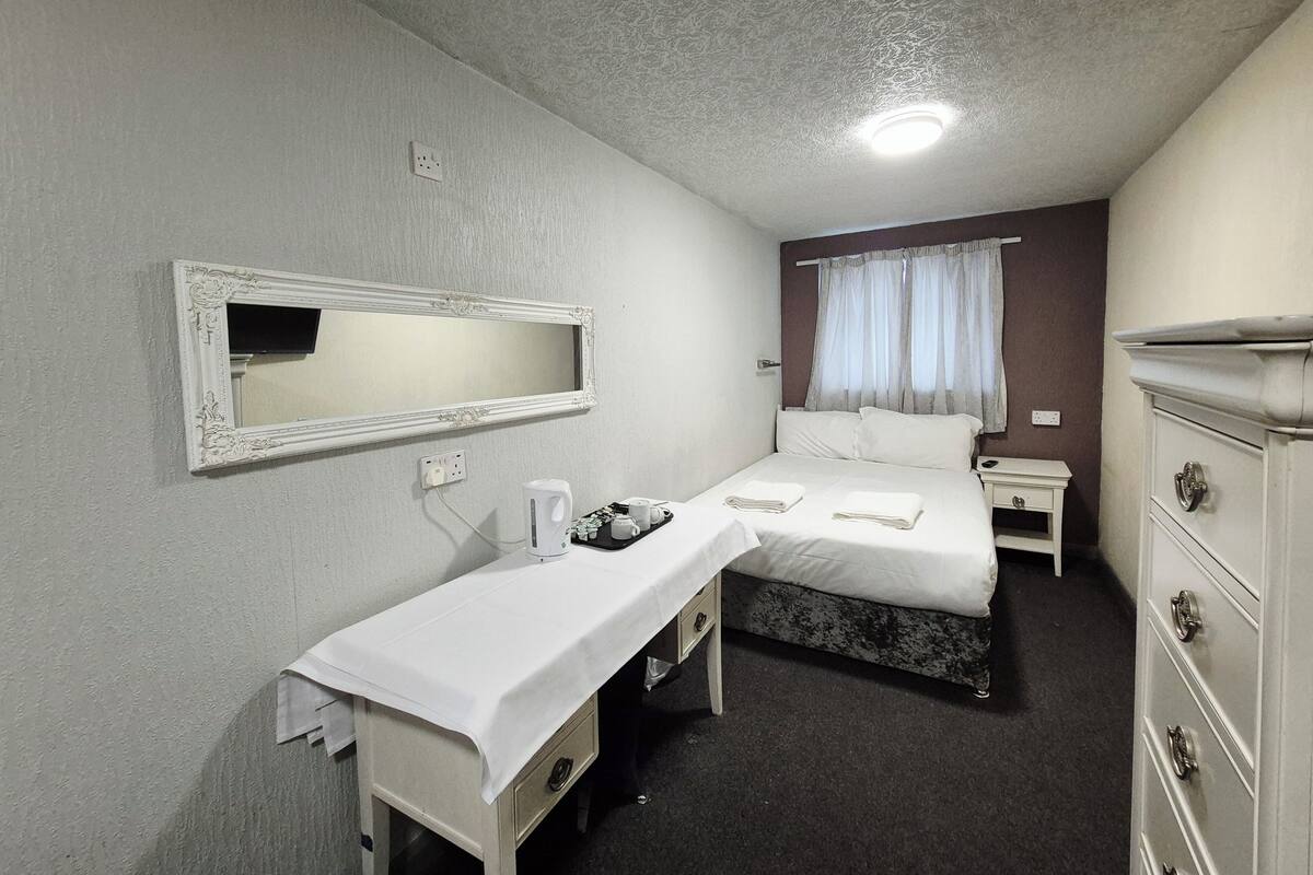 A hotel room with a bed, a bedside table with a mirror, a dresser, and a window with curtains.