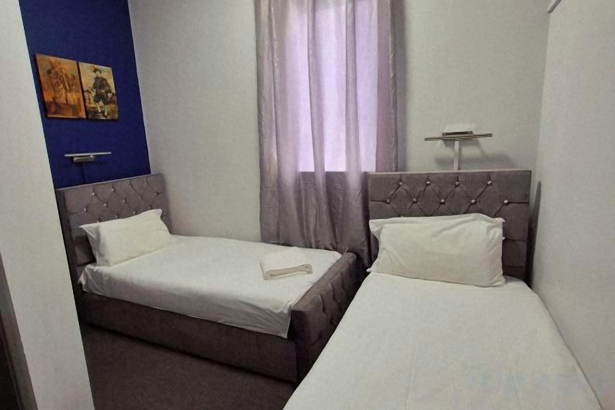 A hotel room with two beds, a nightstand, a wall-mounted lamp, and a window with curtains.