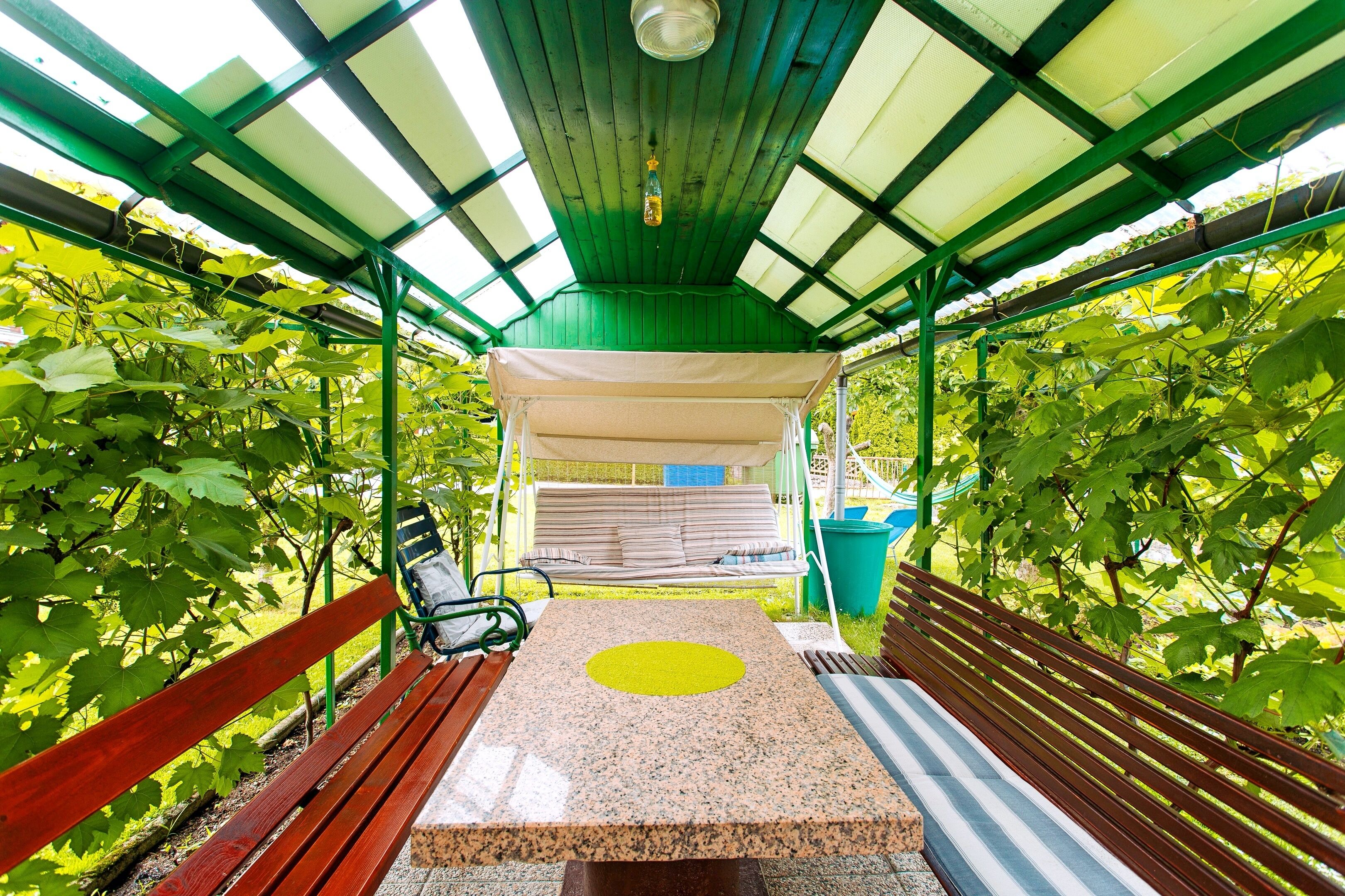 A covered outdoor seating area with benches and a table, surrounded by greenery.