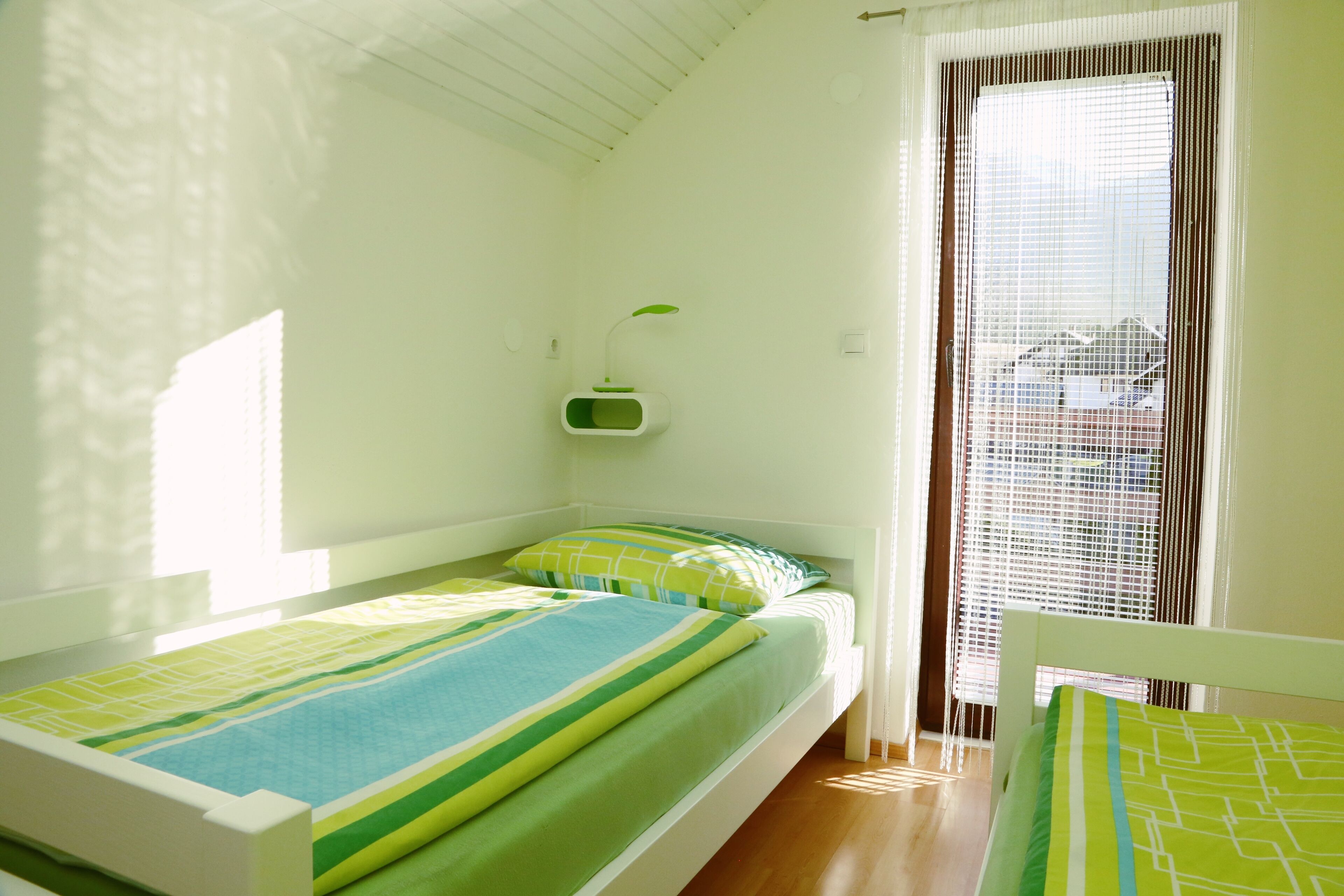 A small, bright room with a single bed, a window with blinds, and a green hanging plant.