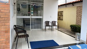 A clean, tiled entrance with a glass door, two wicker chairs, a blue mat, and a metal trash can. - Pousada universitária (Teresina)