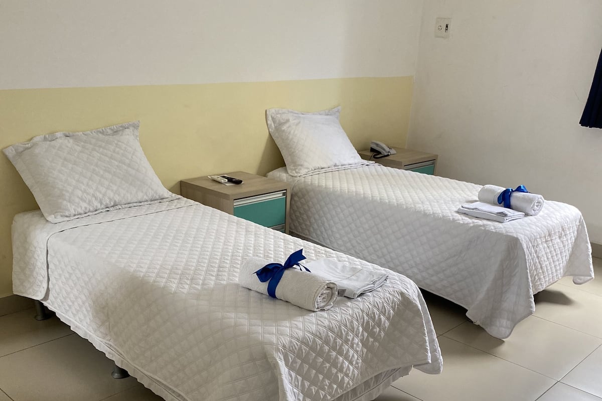 Duas camas de solteiro com roupa de cama branca e detalhes azuis, uma mesinha de cabeceira com telefone e uma tomada na parede.