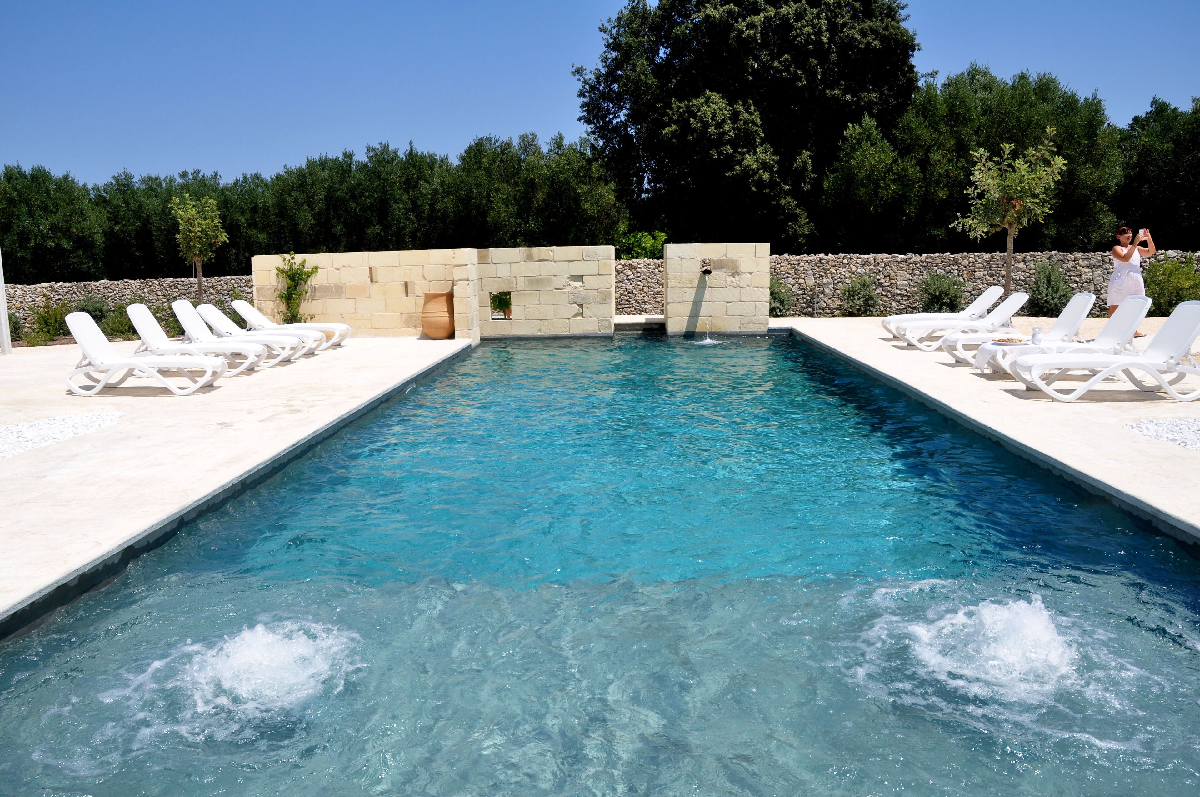 Une piscine rectangulaire à l’eau d’un bleu cristallin, entourée de chaises longues blanches et d’un mur en pierre.