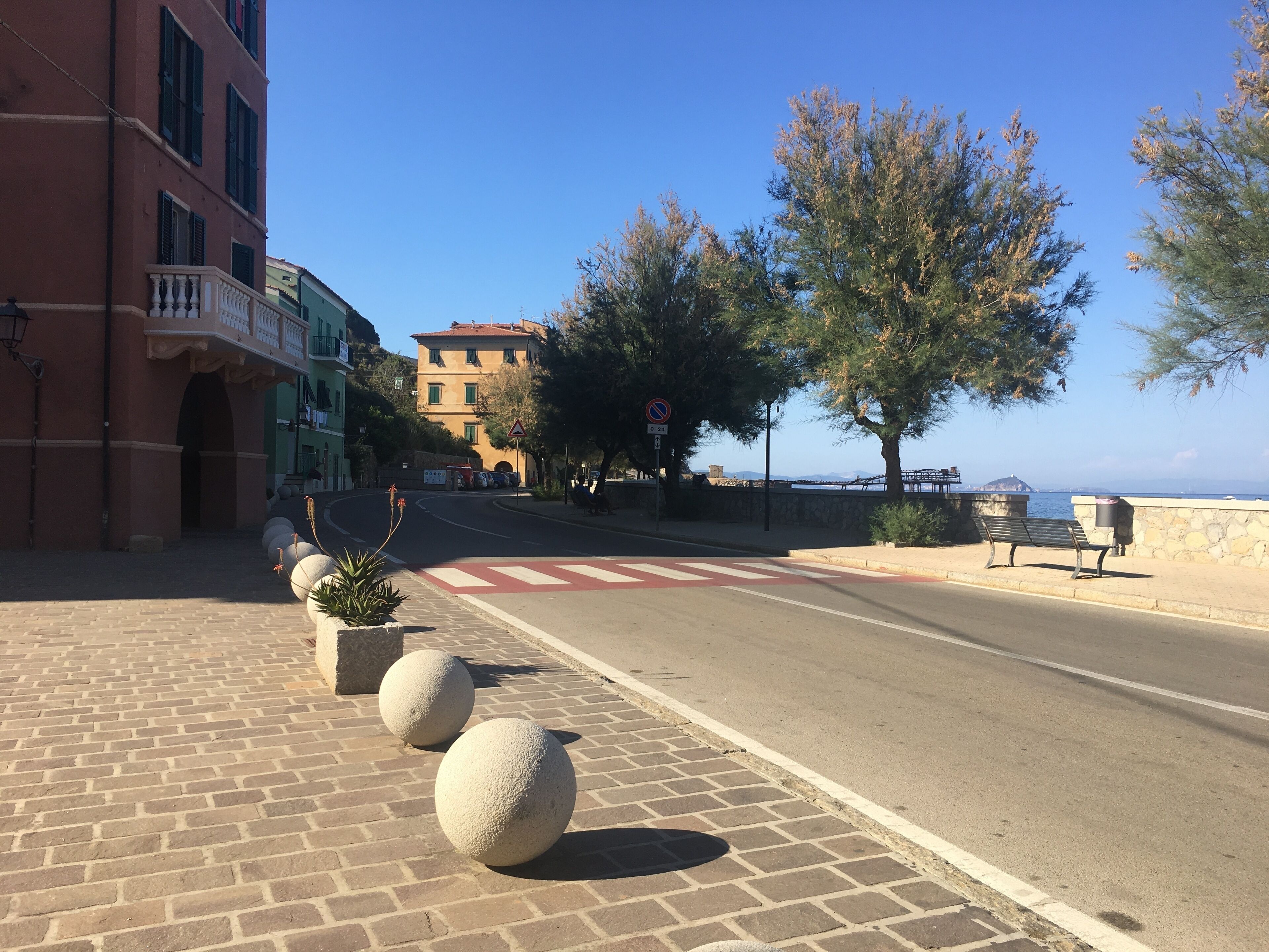 Una calle costera con acera pavimentada, un camino con señal de 'prohibido estacionar' y un banco junto al mar.