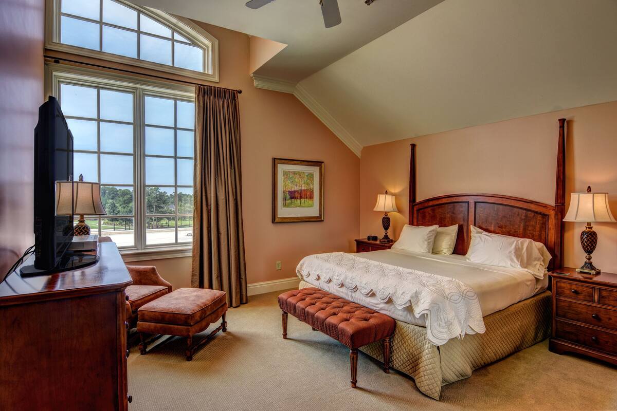 A bedroom with a large bed, a desk with a television, a chair, and a window with curtains.