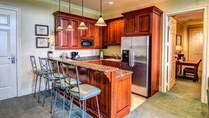 A kitchen with wooden cabinets, a stainless steel refrigerator, a microwave, and a bar area with stools. - The Village at RiverLanding (Wallace)