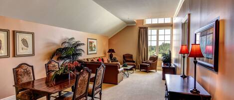 A spacious living room with a high ceiling, a dining area with a wooden table and chairs, and a large window providing natural light.