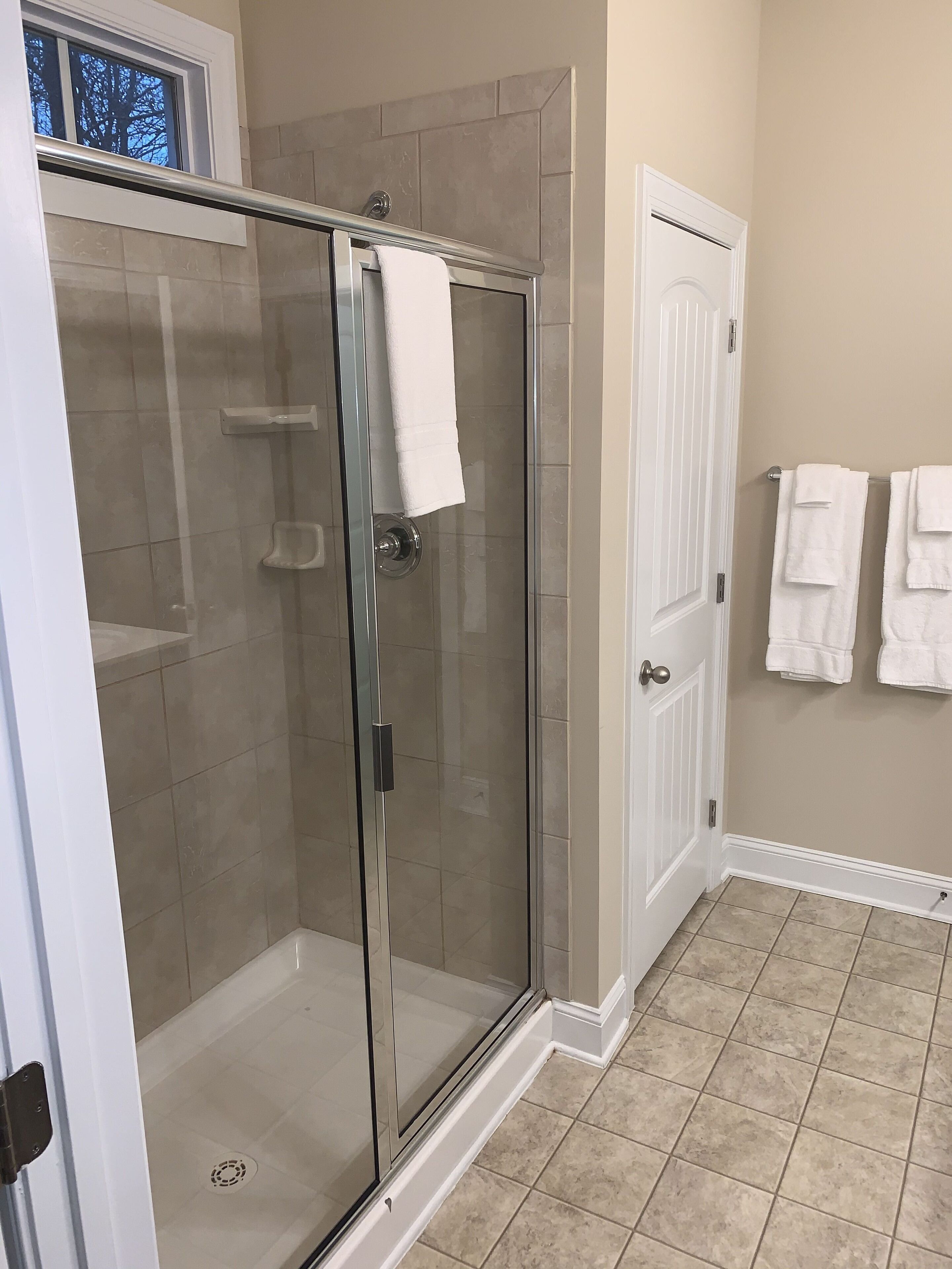 Une douche fermée par des parois de verre, avec une serviette blanche accrochée  à la porte, des murs recouverts de carreaux beiges et une porte blanche ornée de deux serviettes.