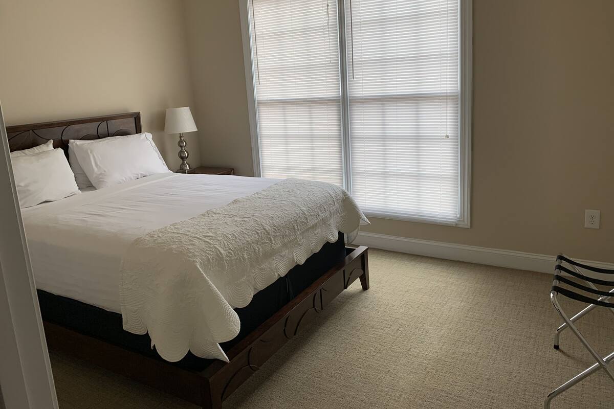 A bedroom with a bed, a nightstand, a lamp, a window with blinds, and a chair.