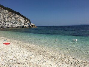A pebble beach with clear water, a red object on the shore, and a rocky cliff. - Small apartment in the center of Rio Marina 10 meters from the sea (Rio Marina)