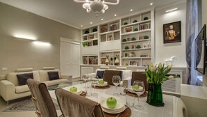 A modern living room with a dining area, featuring a white sofa, a glass coffee table, and a bookshelf filled with decorative items.