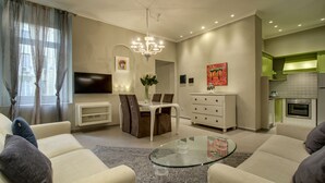 A modern living room with a glass coffee table, beige sofas, a dining area with a white table and chairs, a flat-screen TV, and a kitchen with green cabinets.