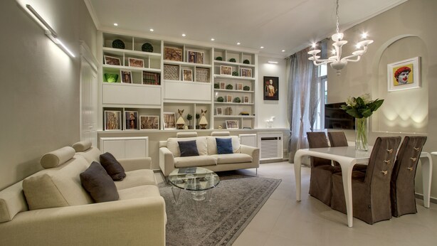 A modern living room with a sofa, coffee table, and dining table. There are shelves with decorative items and a chandelier.