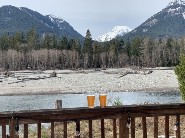 Eine Holzterrasse mit zwei Biergläsern, mit Blick auf einen Fluss, der von Wald und Bergen gesäumt wird.