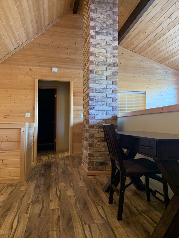 Un interior de madera con un pilar de ladrillo, un escritorio con sillas y una puerta que da a otra habitación.