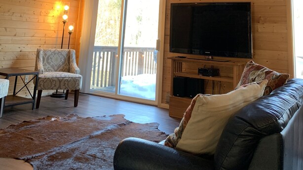 Interior de una cabaña de madera con una televisión grande, un sillón estampado, un sofá de cuero y piso de madera.