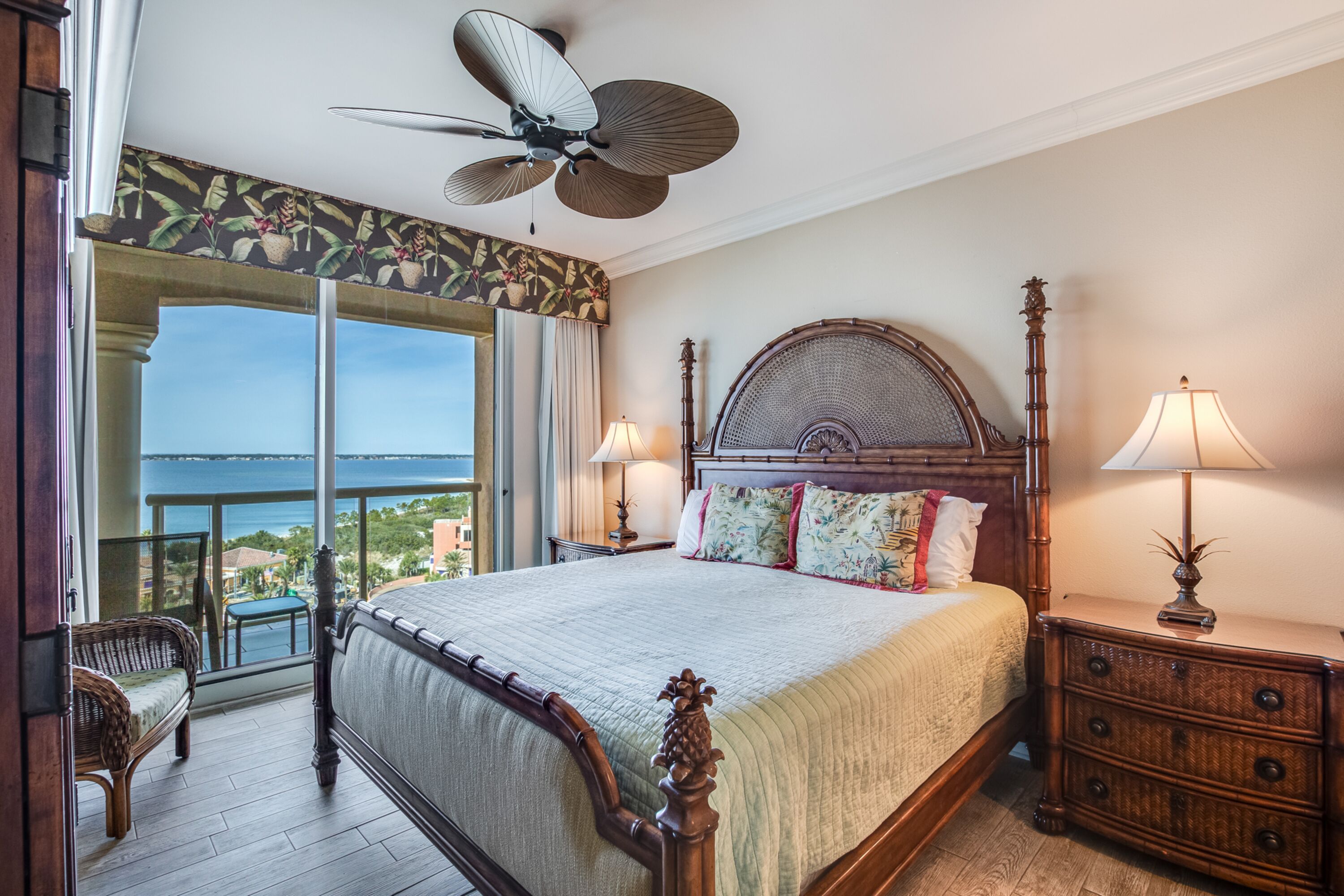 A bedroom with a large bed, a ceiling fan, two bedside lamps, a wooden nightstand, and a view of the ocean through a balcony door.
