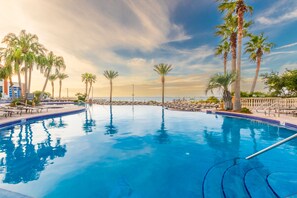 A swimming pool with clear blue water, surrounded by palm trees and lounge chairs, with a view of the ocean in the background. - 1 Portofino 1005 (Pensacola Beach)