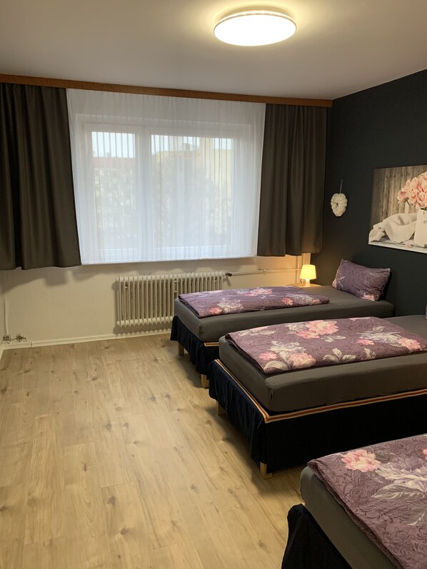 A bedroom with two beds, a radiator, a window with curtains, a lamp, and a wall decoration. - Just Berlin (Bezirk Neukölln)