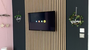 Une chambre moderne avec un mur noir, un téléviseur fixé sur un panneau de lattes en bois, une petite table avec une lampe et des objets décoratifs, notamment un panneau en forme de cœur et une guirlande.