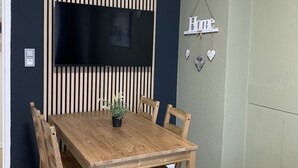 Un coin repas avec une table et des chaises en bois, un téléviseur fixé au mur et des stores verticaux, ainsi qu’une enseigne « Accueil » décorée de cœurs.