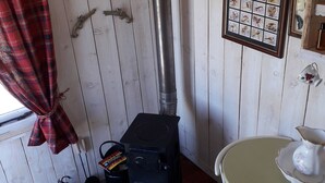 L’intérieur d’un chalet rustique, avec un poêle à bois, des rideaux à carreaux, un tableau représentant un oiseau encadré et une petite table sur laquelle se trouve une carafe.