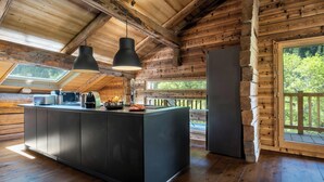 A modern kitchen with a central island, wooden beams, and a large window overlooking greenery. - Chalet Victorina - stunning ski home for 12, close to slopes - OVO Network (La Giettaz)