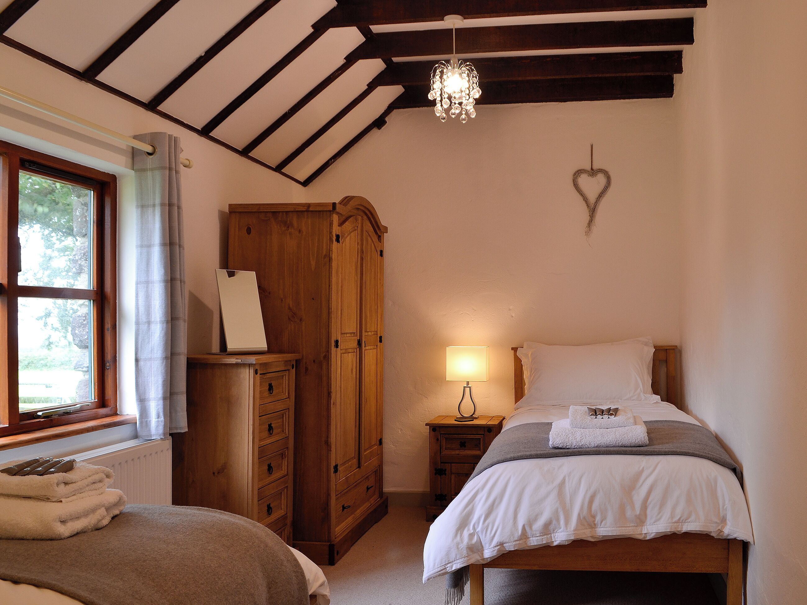 A bedroom with a wooden wardrobe, a bedside table with a lamp, a bed with white bedding, and a chandelier.