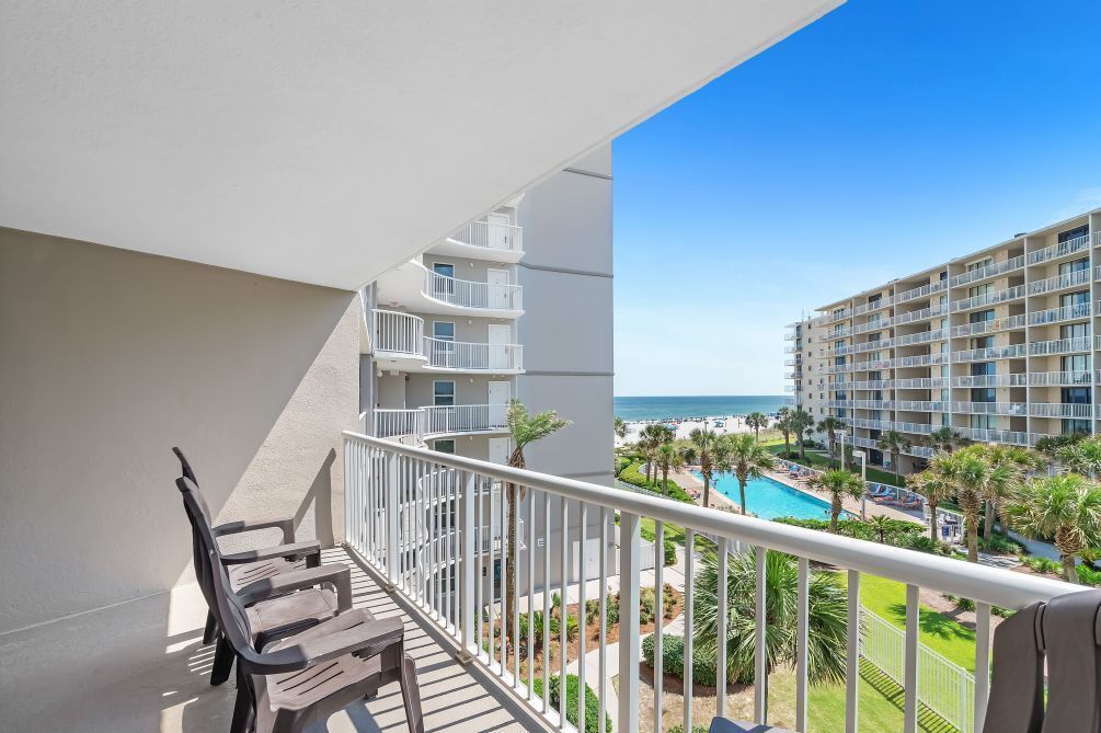 A balcony with wooden chairs, a pool, and a view of a resort area with multiple buildings and palm trees.