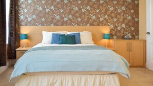Een keurig opgemaakt bed met een blauw-witte dekbedovertrek, twee bedlampen, een houten nachtkastje en een behang met een bloemenpatroon op de muur.