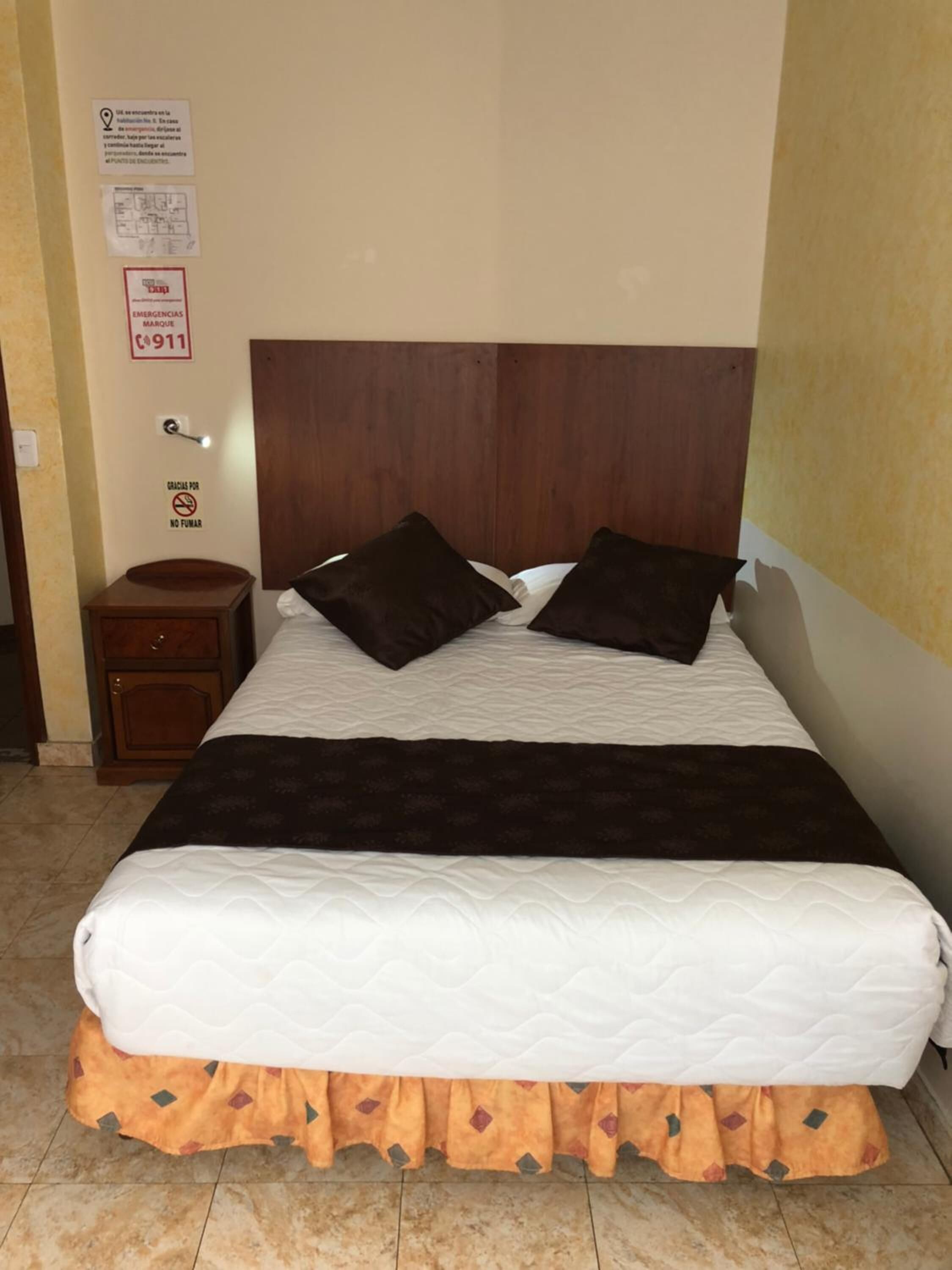 Une chambre d’hôtel avec une tête de lit en bois, un lit avec une literie blanche et marron, deux oreillers, une table de chevet et un panneau indiquant les numéros d’urgence, fixé au mur.