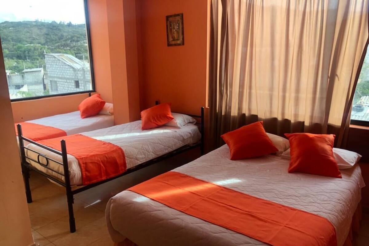 Habitación de hotel con dos camas individuales, cada una con colcha y almohadas anaranjadas. La habitación tiene un ventanal con cortinas, vistas a edificios y suelo de baldosas.