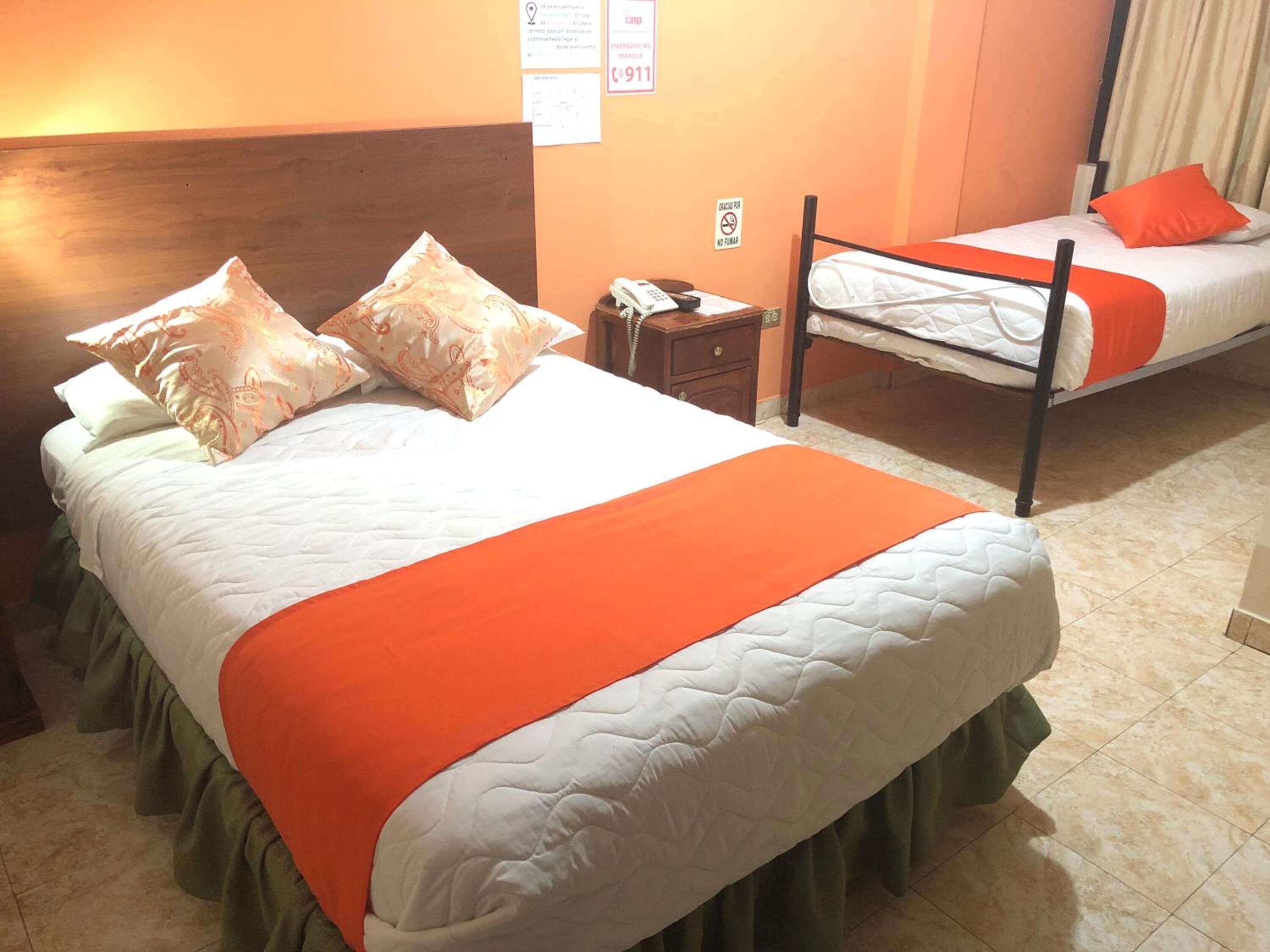 Habitación de hotel con dos camas, cada una con colchón blanco y ropa de cama naranja. Una cama tiene un cabecero de madera a la izquierda, y la otra, una estructura metálica. Se ve una mesita de noche con teléfono entre las camas.