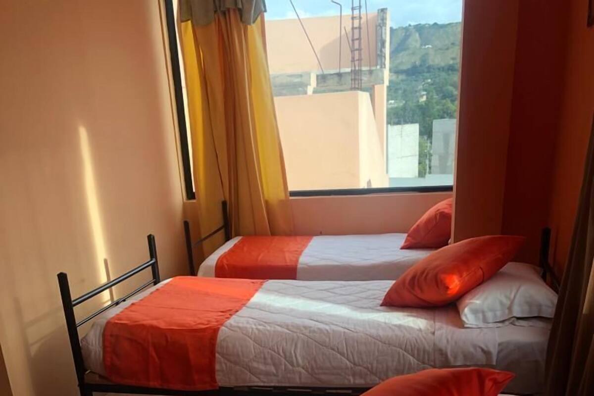 Habitación de hotel con dos camas, cada una con una colcha roja y blanca y almohadas a juego. La habitación tiene una ventana con cortinas, y la vista incluye edificios y un paisaje montañoso.