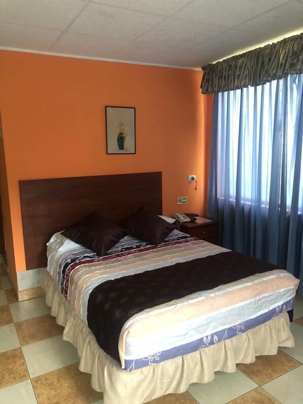 A hotel room with a large bed, orange walls, a framed picture on the wall, and a window with curtains.