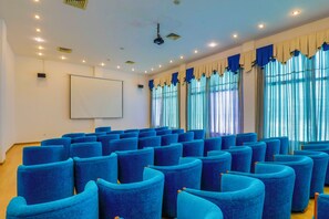 A conference room with rows of blue chairs, a white projection screen, and large windows with curtains.