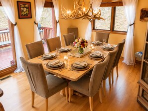 A dining room with a wooden table set for six, surrounded by chairs, a chandelier, and a window with curtains. - Hummingbird Haven - Private Creek (Conifer)