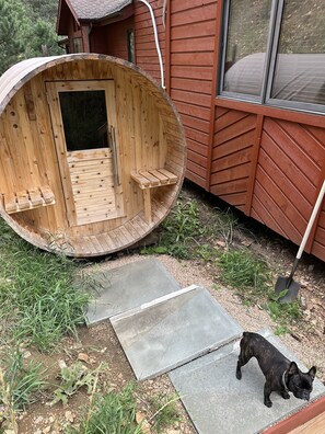 A wooden outdoor hot tub with a door and steps leading up to it, a dog, and a shovel on the ground.