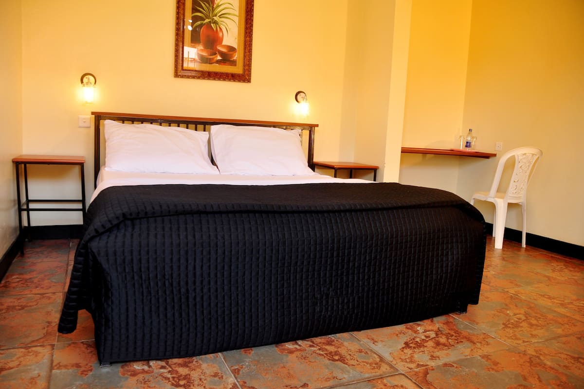 Uma cama bem arrumada com cobertura escura, duas mesas de cabeceira com abajures, uma cadeira branca pequena e uma prateleira fixada na parede com um vaso de planta.