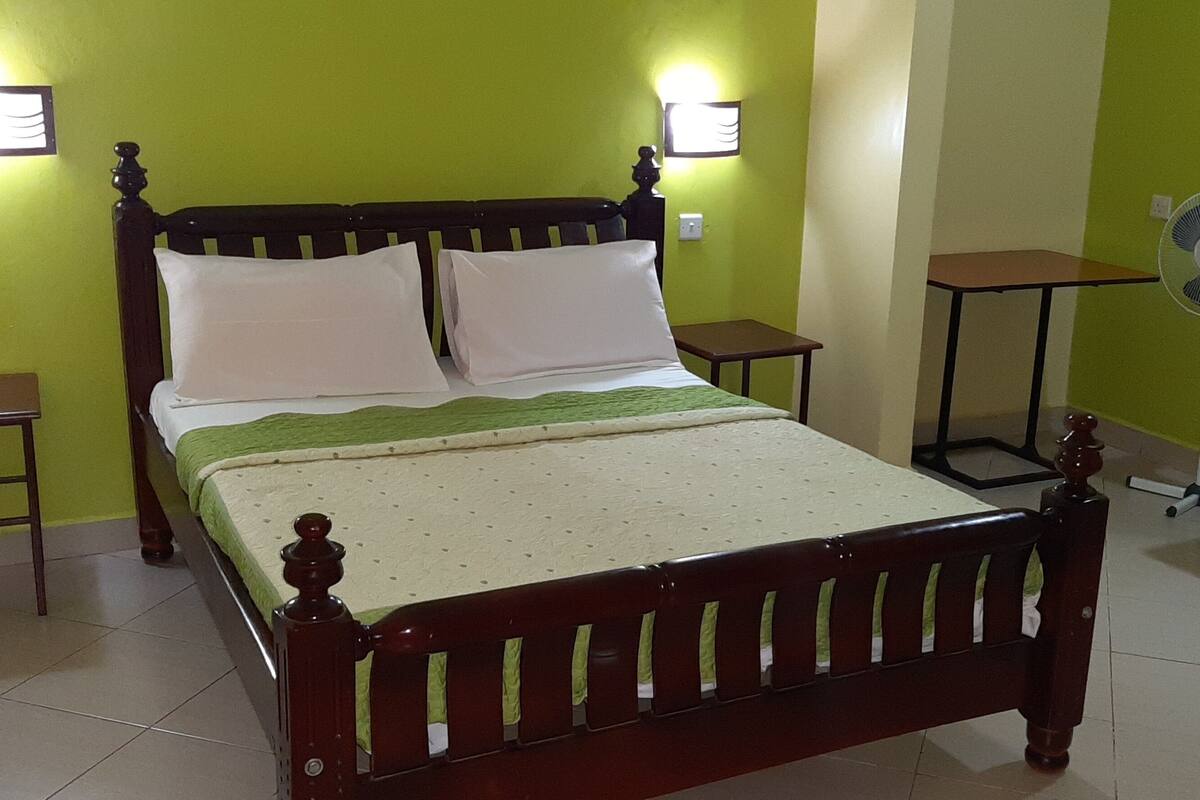 Quarto com cama, duas mesas de cabeceira, um ventilador portátil e uma pequena mesa com uma cadeira.