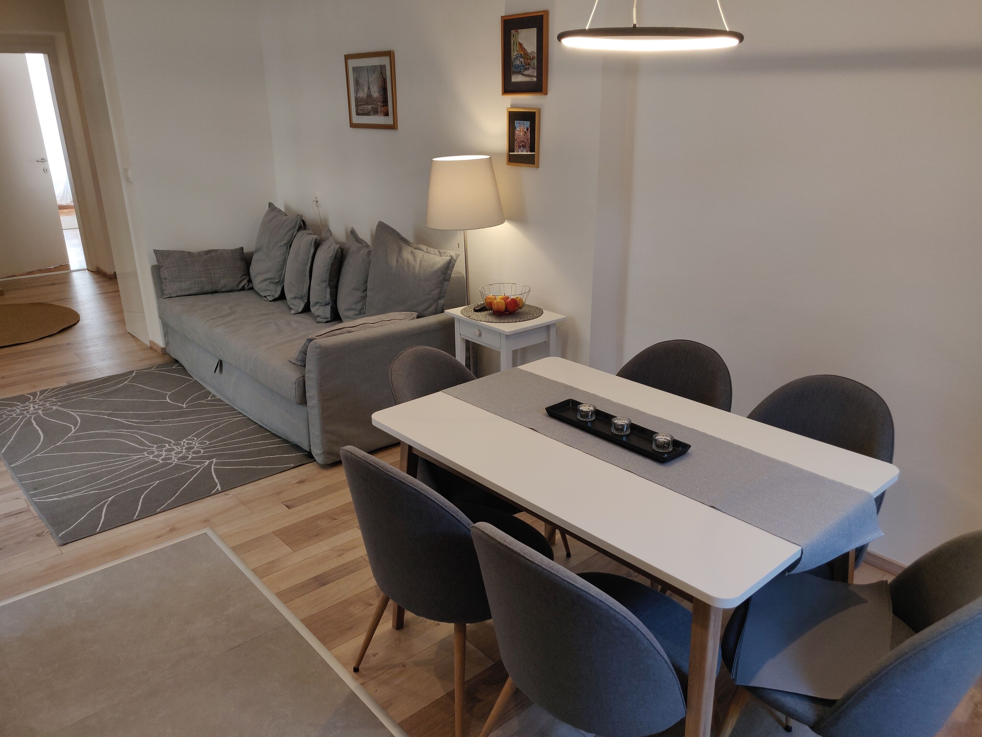 Ett modernt vardagsrum med en grå soffa, ett matbord med fyra stolar, ett litet sidobord med en lampa och en inramad tavla på väggen.