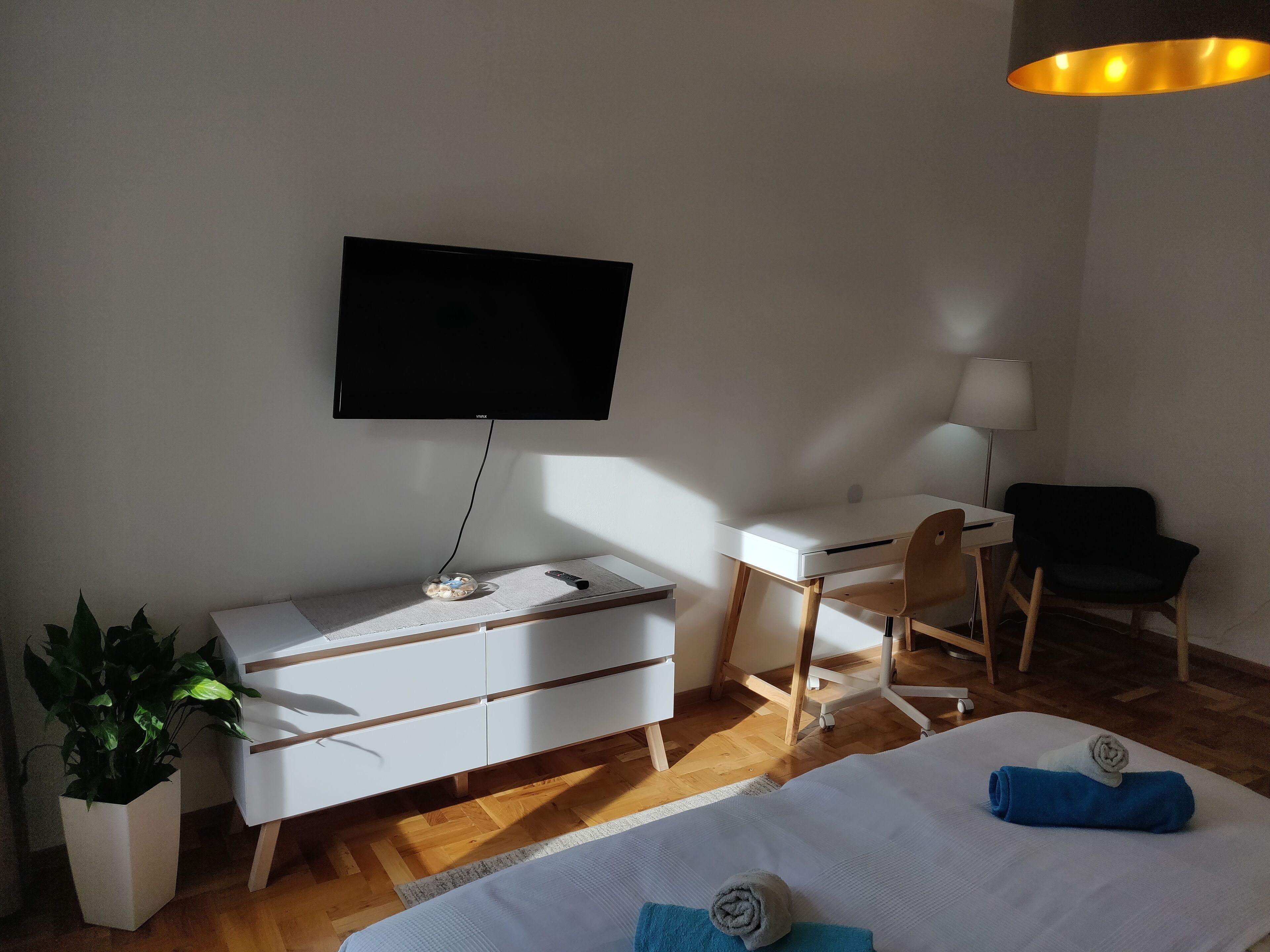 Habitación de hotel moderna con televisión de pantalla plana montada en la pared, un escritorio con silla, cama con edredón blanco y toallas azules, una planta en maceta y suelo de madera.