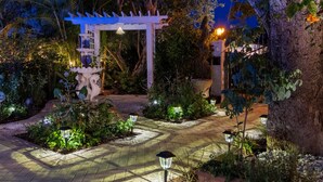 Een goed verlichte tuinpad met een witte pergola, omringd door weelderige begroeiing en sfeervolle verlichting.