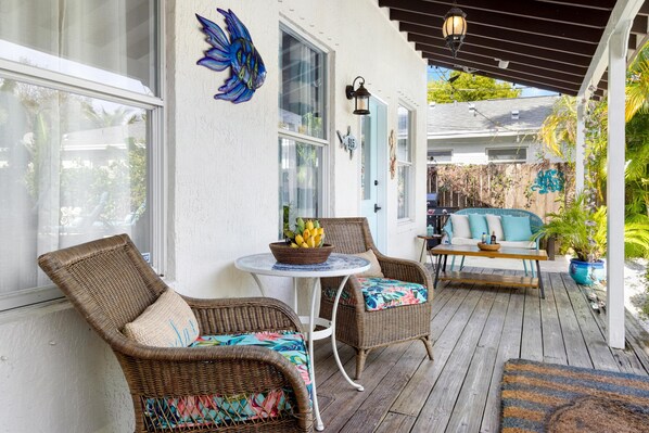 Een overdekte veranda met rieten stoelen, een kleine tafel met een schaal fruit en een blauwe deur.